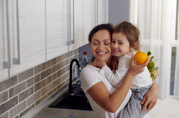 Мать обнимает свою маленькую дочь, стоя на кухне во время приготовления завтрака - Фото, изображение