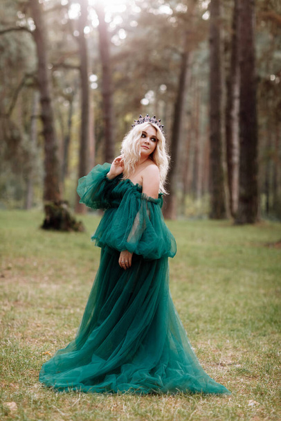 jonge mooie blonde haar vrouw koningin. Prinses loopt. herfst groen bos mystiek. Vintage middeleeuwse glanzende kroon. Lange avond groene jurk. magische fantasie. - Foto, afbeelding