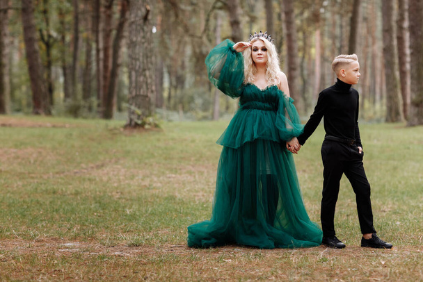 Genç ve güzel sarışın bir kraliçe ve siyah elbiseli genç bir çocuk. Prenses anne oğluyla yürüyor. Sonbahar Yeşil Ormanı Gizemi. Klasik ortaçağ parlak tacı. Uzun akşam yeşil elbisesi. Sihirli fantezi. - Fotoğraf, Görsel