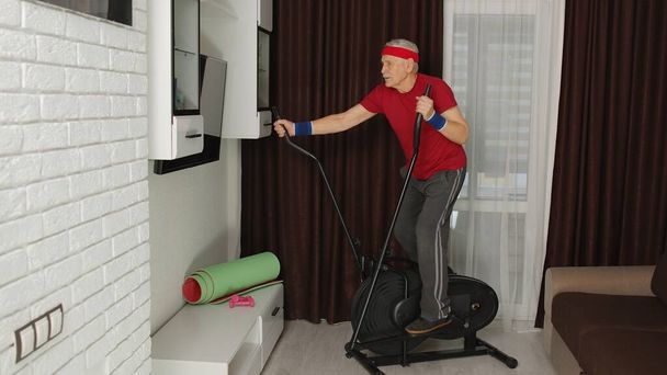 Starszy mężczyzna w odzieży sportowej korzystający z orbitreka w salonie wykonujący ćwiczenia sportowe cardio w domu - Zdjęcie, obraz