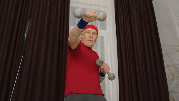Senior oudere man in sportkleding doet gewicht tillen halters workout cardiotraining thuis - Foto, afbeelding
