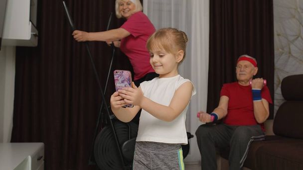 Couple aîné en utilisant orbitrek faire haltères exercices, fille enfant faire selfie sur téléphone mobile - Photo, image