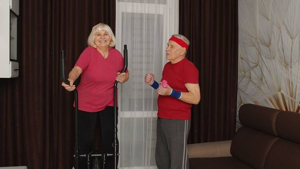 Old senior couple exercising on orbitrek, doing weight lifting sport dumbbells exercises at home - Photo, Image