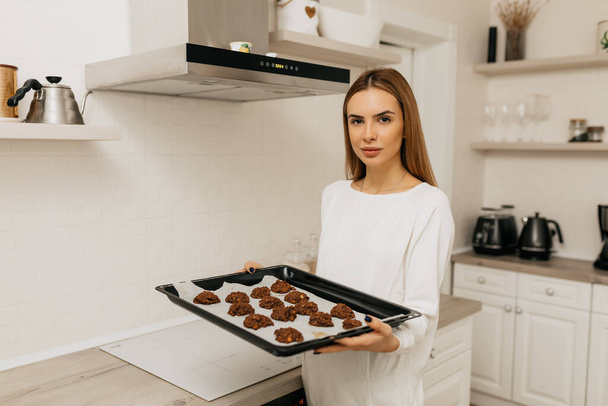 Jovem senhora com cabelo castanho claro em camisa branca cozinhar um biscoitos de chocolate caseiros na cozinha leve. Foto de alta qualidade - Foto, Imagem