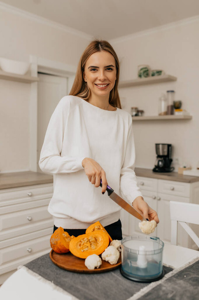 Mutfakta sebzelerle yemek pişirirken kameraya poz veren beyaz gömlekli mutlu genç bayan. Yüksek kalite fotoğraf - Fotoğraf, Görsel