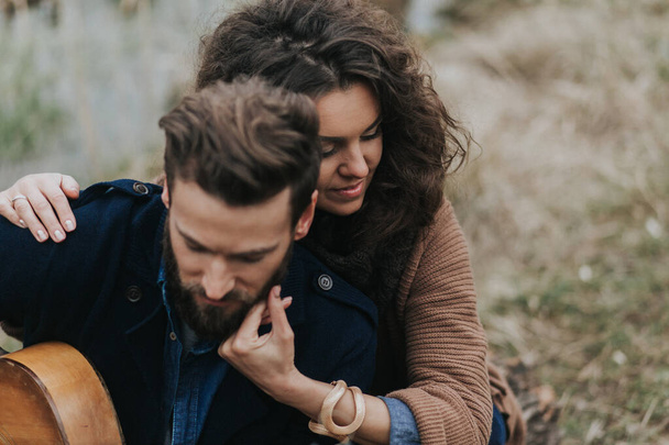 caucasian man is playing guitar with woman by the lake. Young couple is hugging on autumn day outdoors. A bearded man and curly woman in love. Valentine's Day. Concept of love and family - Photo, Image