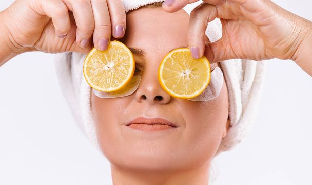 An enlarged portrait of a caucasin elderly woman with lemon slices in front of her eyes on a white background. Skinimalism and senior healthy skin care concept. - Valokuva, kuva