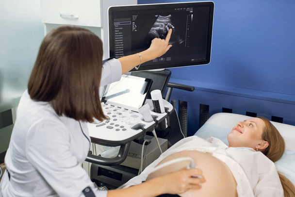 Young excited pregnant woman getting ultrasound in modern clinic, happy to see her baby on the screen. Female doctor pointing on ultrasound of belly of pregnant woman in clinic - Φωτογραφία, εικόνα