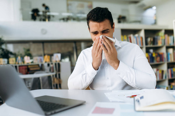 Unhealthy male employee sit at the desk, using a paper tissue, sneezing. Business man feeling unwell, struggling with running nose. Hispanic guy remotely working or studying, while sickness - Photo, image