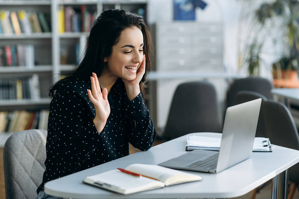 Joyful young woman sit at the work desk, waves hand, greeting colleagues. Attractive business female communicate with colleagues or friends by video call, smiling, remotely work or study - Photo, Image