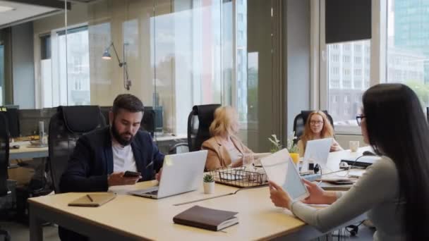 Közepes felvétel barátságos, többnemzetiségű munkatársak csoportjáról, akik együtt ülnek a közös asztalnál, dolgoznak és beszélgetnek a modern, nagy ablakokkal rendelkező, nyílt téri irodában. - Felvétel, videó