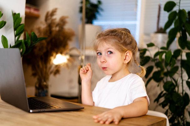 Egy kislány ül egy asztalnál egy laptoppal feltartott ujjal, mosolyog és okos lesz, boldogságot tapasztalva.. - Fotó, kép