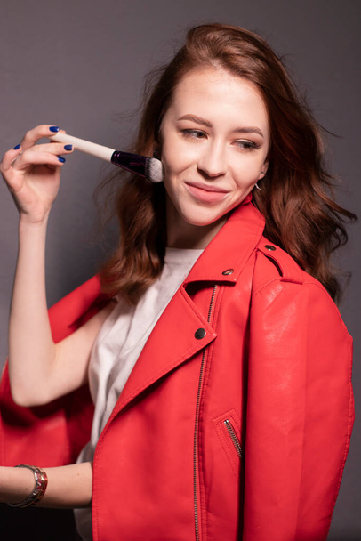 charmante brunette vrouw in rood leren jas met make-up borstels geïsoleerd op grijze muur achtergrond. professionele make-up artiest. kopieerruimte - Foto, afbeelding