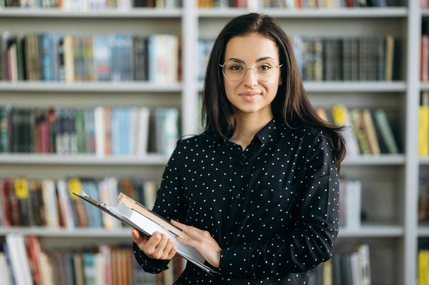 Portret van een aantrekkelijke zelfverzekerde jonge vrouw, student, leraar of zakenvrouw, in bril, kijkt direct naar de camera met boeken in haar handen, staand op de achtergrond van boeken - Foto, afbeelding