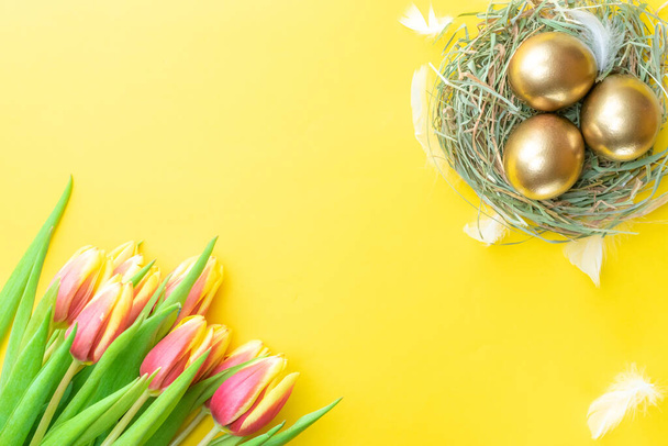 Easter eggs gold. Golden colour egg in basket with spring tulips, white feathers on pastel yellow background in Happy Easter decoration. Congratulatory easter design. Flat lay, top view - Photo, Image