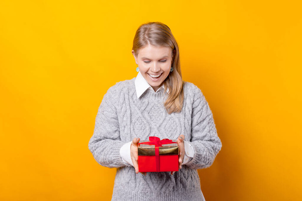 Sorprendida joven está sosteniendo un regalo que tiene sobre fondo amarillo. - Foto, Imagen