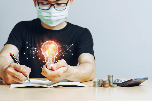 Молодые азиатские мужчины держат лампочку в маске, работая из дома с идеями новых идей, инновационных технологий для исследований данных и монет на деревянном столе в офисе.. - Фото, изображение