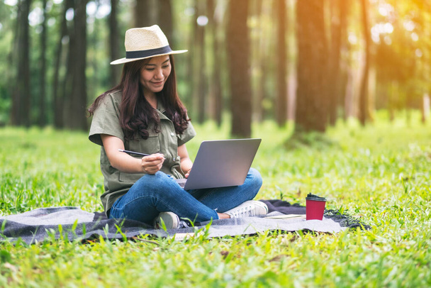Egy gyönyörű ázsiai nő dolgozik és gépel a laptop billentyűzetén, miközben ül a parkban - Fotó, kép