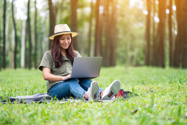 Een mooie aziatische vrouw werken en typen op laptop toetsenbord terwijl zitten in het park - Foto, afbeelding