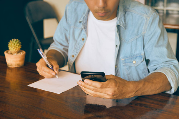Young Asian Casual Businessman in Denim or Jeans Shirt Using Smartphone and Take Note in Coffee Shop. Casual Businessman Working with Technology in Vintage Tone - Photo, Image