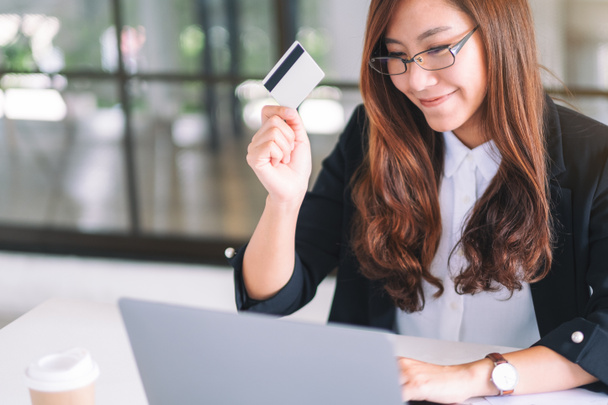 Una mujer de negocios asiática sosteniendo tarjetas de crédito mientras usa computadora portátil en la oficina - Foto, Imagen