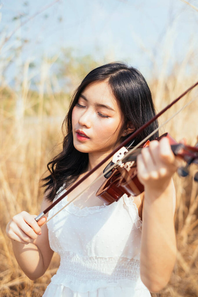 Czarne długie włosy kobieta w białej sukience grać na skrzypcach w trawie w wietrzny dzień. - Zdjęcie, obraz