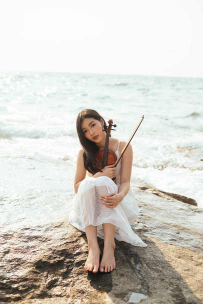 Красива чорна довге волосся жінка в білій сукні сидить з колінами вгору і обіймає скрипку на скелі
. - Фото, зображення