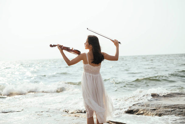 Back shot of white dress woman playing violin at the sea with wave. - Foto, imagen