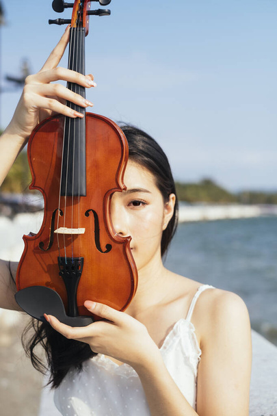 Black hair woman in white dress raise her violin at her face while standing in the sunshine. - Photo, Image