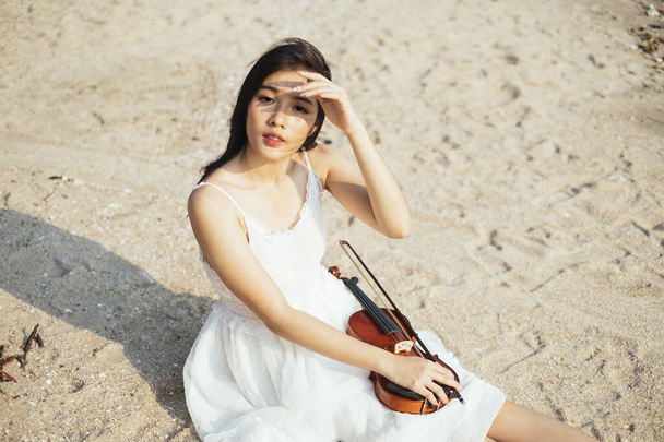 Violinista negra de pelo largo sentada en la playa de arena con violín en sus rezagos y levantando la mano para proteger sus ojos de la luz del sol. - Foto, Imagen