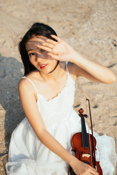 Černá dlouhosrstá houslistka sedící na písečné pláži s houslemi na vlasech a pozvedá ruku, aby si chránila oči před slunečním světlem, vertikálně. - Fotografie, Obrázek