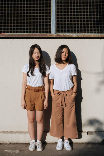 Két lány fehér pólóban és barna nadrágban, együtt állnak a napfényben a falnál, fogják egymás kezét, teljes erővel.. - Fotó, kép