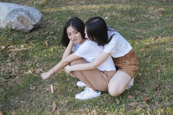 Két lány fehér pólóban és barna nadrágban ül a füvön, hosszú har lány ölel rövid hajú lányt hátulról és mosolyognak együtt.. - Fotó, kép