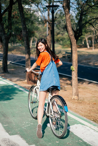 Uzun saçlı, turuncu tişörtlü ve kot pantolonlu bir kızın parkta bisiklet yolunda bisiklet sürüşünün arka planı.. - Fotoğraf, Görsel
