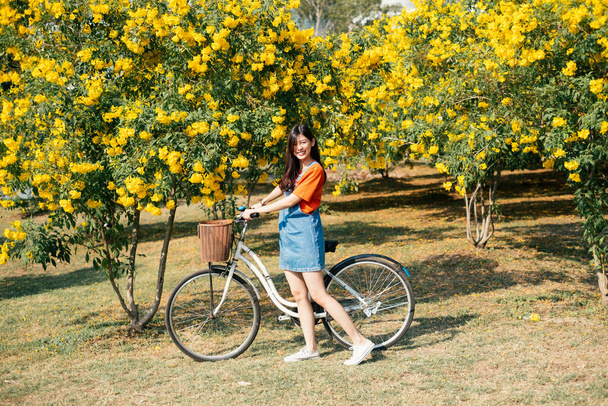 Menina de cabelo longo em camiseta laranja e calça jeans em pé no jardim de flores amarelas com bicicleta. - Foto, Imagem