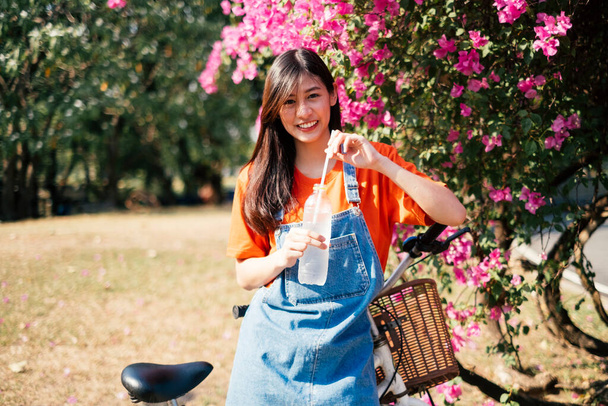 Long hair girl in orange t-shirt and jeans jumper standing at the pink bougainvillea with her bike and holding the drinking water in her hand. - Foto, Imagem