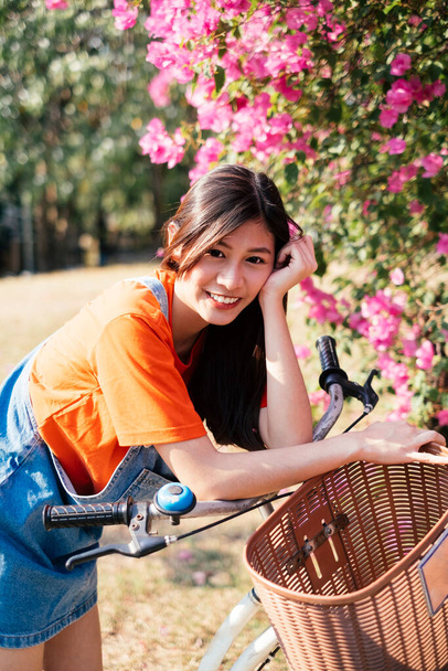 Menina de cabelo longo em camiseta laranja e jeans jumper parar a bicicleta no bougainvillea rosa e dobrar o corpo na bicicleta. - Foto, Imagem