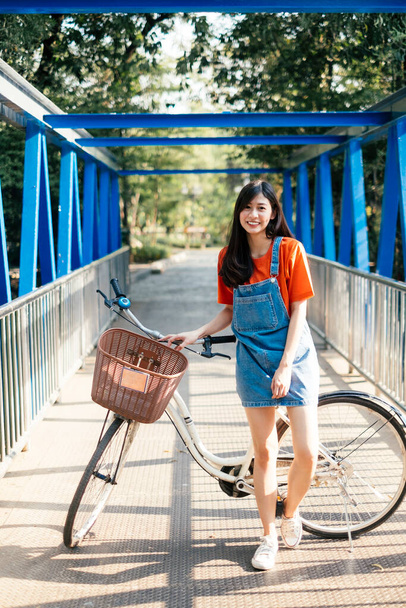 Девушка с длинными волосами в оранжевой футболке и джинсах, стоящая со своим велосипедом на голубом мосту. - Фото, изображение