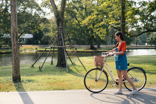 Turuncu tişörtlü ve kot pantolonlu uzun saçlı kız bisikletle parkta çimen tarlasında yürüyor.. - Fotoğraf, Görsel