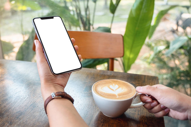 Mockup image of a woman holding and using black mobile phone with blank desktop screen while drinking coffee in cafe - Valokuva, kuva
