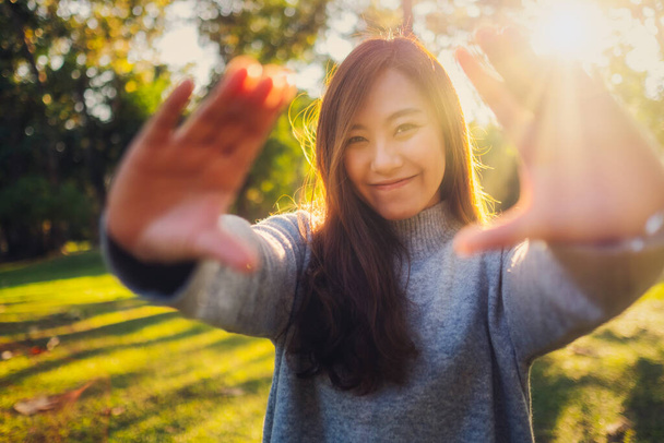 Portrait image of a beautiful asian woman raising hands and playing with camera in park before sunset - Photo, Image