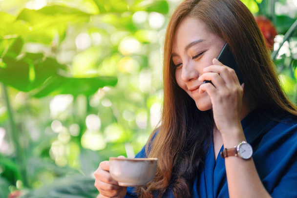 Крупним планом зображення азіатки, що тримає і розмовляє на мобільному телефоні під час пиття кави в саду
 - Фото, зображення
