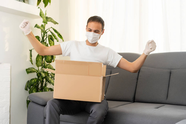 пост, дом и образ жизни - человек с коробками в маске и перчатками на диване - Фото, изображение
