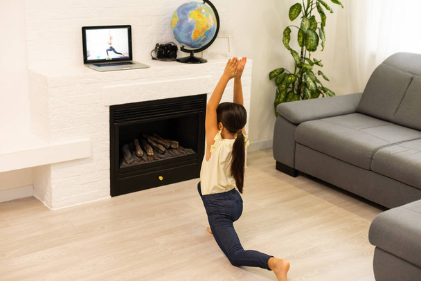 Little girl watching online video on laptop and doing workout or fitness exercises in her room at home. Distant online training, social distance self-isolation, online education concept - Photo, Image