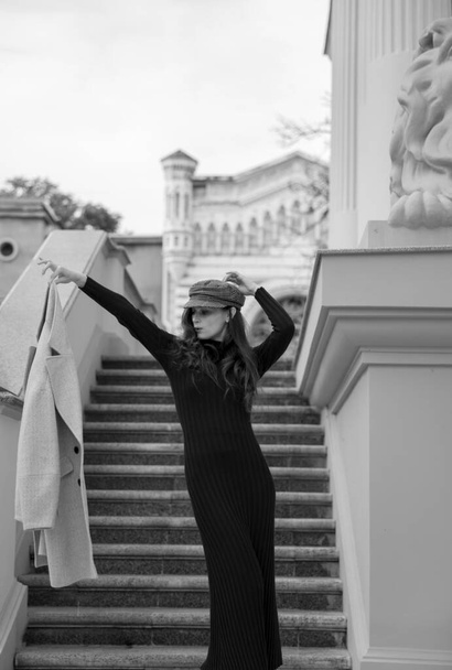Fekete-fehér kép egy női modellről, szűk fekete ruhában, flörtöl és a kabátját a kezében tartja.. - Fotó, kép