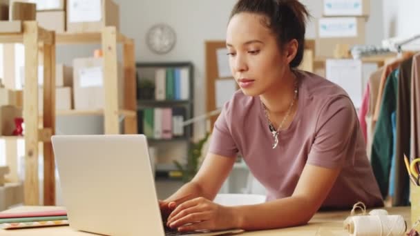 Fiatal vegyes fajú nő dolgozik laptop és jegyzetel az íróasztal irodájában az online ruhaüzlet - Felvétel, videó