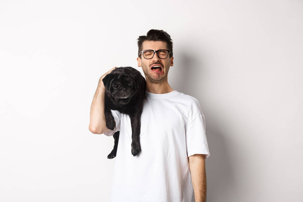 Smutny właściciel psa płacze, trzymając ładny czarny mops na ramieniu i wyglądając nędznie, szlochając stojąc nad białym tłem - Zdjęcie, obraz