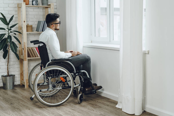 Young disabled man sitting in a wheelchair near the window - Φωτογραφία, εικόνα