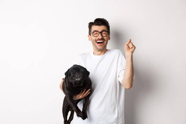 Cheerful handsome man with dog rejoicing, celebrating victory. Happy guy carry cute black pug and looking upbeat, adopting pet, standing over white background - Foto, imagen