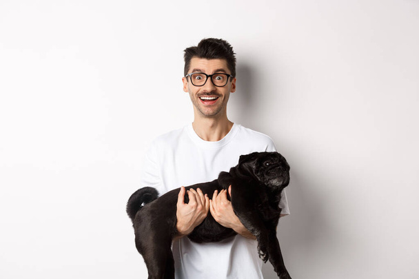 Happy hipster in glasses holding cute black pug and smiling, dog owner staring at camera with amazed smile, standing over white background - Foto, imagen
